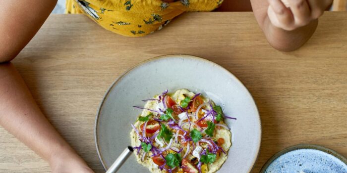 Natural overhead shot of beautiful brunette ethnic vegan female in a bright yellow floral dress eating tasty fine, organic, international cuisine on artisanal ceramic plate including naan bread, tomatoes and herbs drinking a red vegetable juice from a reusable metal straw.