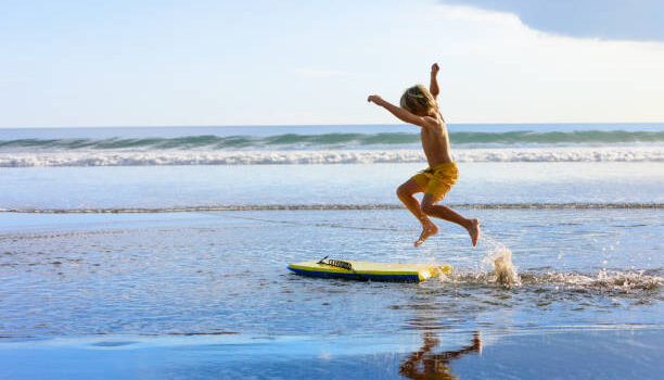Happy boy - young surfer with bodyboard have fun on beach, run by sea water pool. Active family lifestyle, kids outdoor water sports, swimming activity in surf camp. Summer vacation with child.