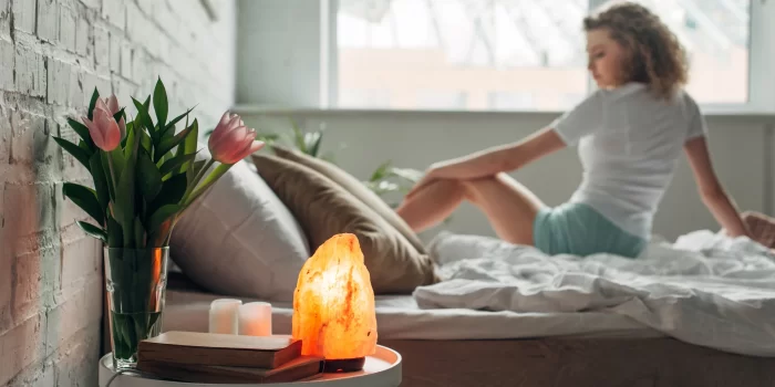 beautiful girl on bed with Himalayan salt lamp, flowers and books on table in bedroom, selective focus